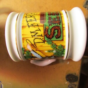 May include: A white ceramic shaving mug with gold trim and a yellow and green graphic. The graphic features the text "J.M. FERRER" and an illustration of a person holding a rope. The mug has a handle and a decorative design.