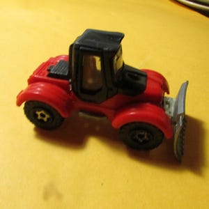 May include: A small red and black toy tractor featuring a front-mounted silver plow blade. The vehicle has a black cabin, black wheels with star-shaped centers, and a red body. The design includes a detailed cab structure and a functional-looking blade.