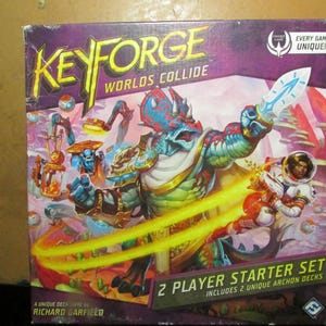 May include: KeyForge Worlds Collide 2 Player Starter Set board game box featuring a large blue and red dinosaur-like creature holding a glowing blade, alongside a space-suited figure, with the text KeyForge Worlds Collide and Richard Garfield.