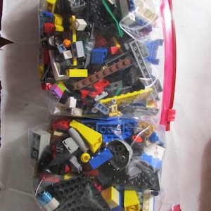 May include: Two clear plastic bags filled with colorful toy building blocks. The blocks are various sizes and shapes, including bricks, plates, and specialty pieces in yellow, blue, red, black, and white. The bags are sealed with a red zipper.