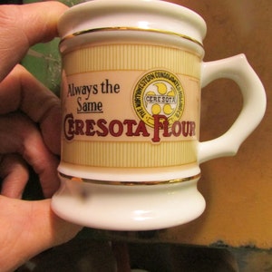 May include: A small, white ceramic mug with a handle, featuring the text "Always the Same CRESOTA FLOUR." The mug has gold trim and a cream-colored background with vertical lines. The mug also has a logo that says "The Northwestern Consolidated Milling Co."