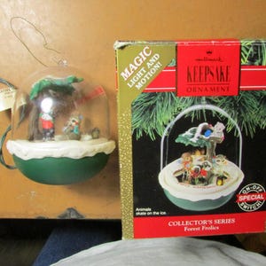 May include: A Hallmark Keepsake ornament featuring a miniature forest scene inside a clear dome. The ornament has a green base and a white trim. The box reads "Magic Light and Motion!" and "Collector's Series Forest Frolics."