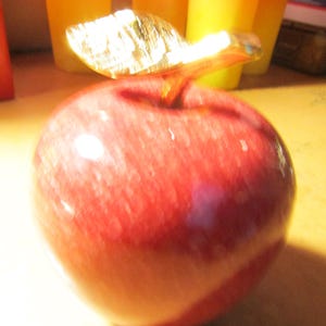 May include: A red apple-shaped decorative object with a gold-colored leaf detail. The apple has a glossy finish and is likely made of glass or a similar material. The background includes blurred orange and yellow objects.