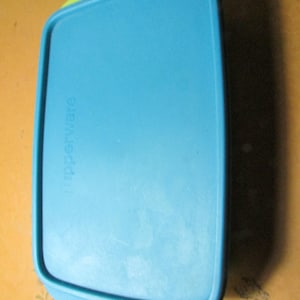 May include: A rectangular, blue Tupperware container with a light green rim. The lid has a slightly curved design and the word "Tupperware" is printed on the side. The container is made of plastic.