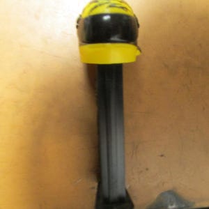 May include: A yellow and black PEZ dispenser shaped like a racing helmet. The helmet has a black visor and a yellow top with black accents. The dispenser's stem is black.