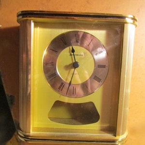 May include: A rectangular gold-toned quartz clock with a light yellow face and a clear glass cover. The clock face has Roman numerals and the word "Benchmark" is printed on the face. The clock has a small, curved opening at the bottom.