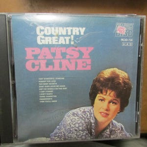 May include: A Patsy Cline Country Great! compact disc in a clear plastic jewel case. The cover features a blue background with white and pink text, a tracklist, and a photograph of Patsy Cline wearing a blue and white patterned shirt.