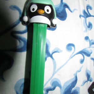 May include: A green and black penguin PEZ dispenser. The penguin has a green hat with a white pom-pom, white eyes, and an orange beak. The dispenser is green and has a rectangular shape.