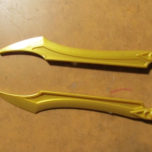 May include: Two gold-colored plastic decorative blades with curved edges and ornate detailing. The blades have a pointed tip and a decorative element at the handle end. The items are likely for cosplay or costume use.