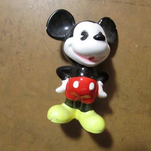 May include: A ceramic figurine of Mickey Mouse, featuring black ears, a white face, and a red shorts. The figure has yellow shoes and white gloves, with a smiling expression. A classic Disney collectible.