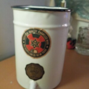 May include: White enamel water dispenser with a black rim and a spigot. The dispenser features two circular labels with text and a red and black design. A vintage-style kitchenware item.