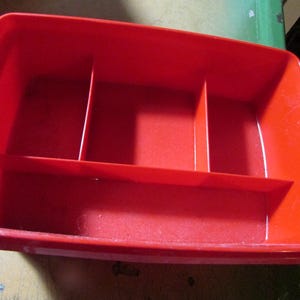 May include: A rectangular red plastic storage tray featuring a single horizontal divider and two vertical dividers, creating four distinct compartments within the container. The tray has smooth, rounded corners and a solid, uniform red color.