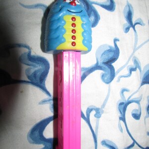 May include: A vintage PEZ dispenser featuring a blue, smiling caterpillar character with red antennae and yellow accents. The dispenser has a pink stem and foot. The background is a white fabric with blue floral patterns.