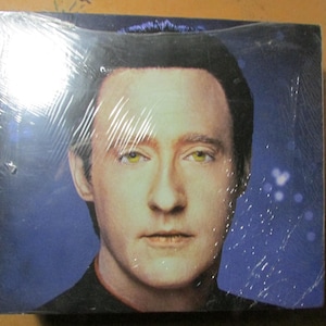 May include: Square package wrapped in clear plastic featuring a portrait of a man with fair skin, dark hair, and yellow-green eyes. The background is a gradient of blue with white star-like shapes.