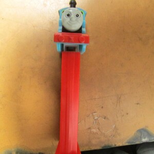 May include: A blue, red, and gray Thomas the Tank Engine PEZ dispenser. The dispenser features a smiling Thomas face on top of a red rectangular base and stem. The stem ends in a heart-shaped design.