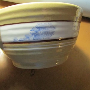 May include: A handmade ceramic bowl with a handle, featuring a beige exterior with brown and white bands. The bowl has a blue floral design on the white band. The bowl is held by a hand.