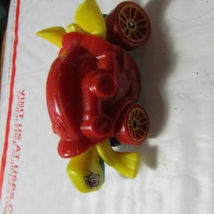 May include: A red and yellow toy car with dark red wheels. The toy car has a unique design with yellow wings and accents. The wheels have a detailed spoke design. The toy is on a white surface.