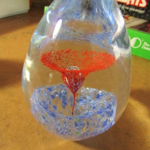 May include: A clear glass paperweight with a red flower design suspended inside. The flower has a textured, dotted appearance. The base of the paperweight features blue and white swirls. The paperweight is egg-shaped.