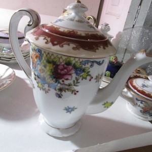 May include: White porcelain teapot with a floral design. The teapot features a burgundy and gold rim, a curved handle, and a decorative lid. The body of the teapot is adorned with colorful flowers and foliage. A classic piece for tea service.