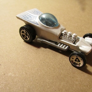 May include: A white toy race car with a clear bubble top and blue flame decals. The car has black wheels with silver rims and a detailed engine. A classic collectible toy.