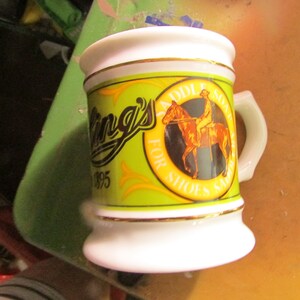 May include: A white ceramic mug with a handle, featuring a green and yellow label with the text "King's 1895" and an illustration of a person on a horse. The mug has gold trim.