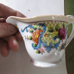 May include: A small, white ceramic creamer with a floral design. The creamer features a handle and spout, and is decorated with orange, blue, and purple flowers. The rim has a gold trim. The creamer is held by a hand.