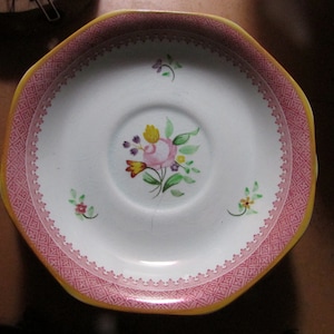 May include: White ceramic saucer with a pink and yellow scalloped edge. The saucer features a pink patterned border and a central floral design with pink, yellow, and green colors. Small floral accents are placed around the edge.