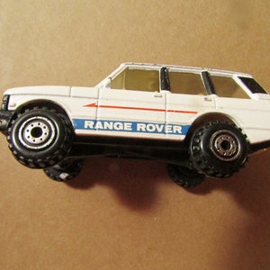 May include: A white Range Rover toy car with black wheels and a blue and red stripe. The toy car has the words "RANGE ROVER" printed on the side. The toy car is a miniature replica of the real vehicle.