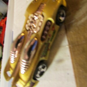 May include: A gold-colored toy car with black wheels and silver accents. The car has a unique design with a streamlined body and decorative elements. The toy car is likely a collectible or a model car.