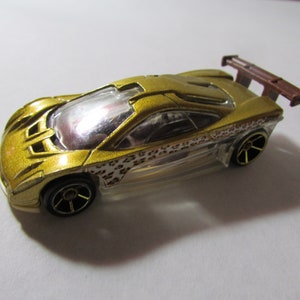 May include: A gold toy race car with a clear windshield and a brown spoiler. The car has black wheels with gold accents and a leopard print design along the side. The car is a miniature replica.