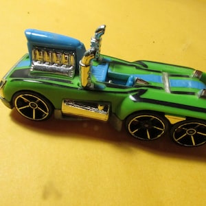 May include: A lime green toy truck with a blue stripe down the center, featuring a large chrome engine block, dual chrome exhaust stacks, and six black wheels with silver rims. The vehicle has a low-profile design with black accent lines.
