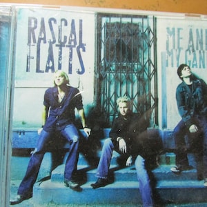 May include: A Rascal Flatts Me and My Gang music CD in a clear plastic jewel case. The cover features three individuals sitting on blue steps in front of a metal gate, with the band name and album title printed in blue text against a light background.