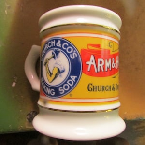 May include: A small, white ceramic mug with a handle, featuring the "Church & Co's Baking Soda" logo in a blue circle and the "Arm & Hammer" logo in red on a yellow background. The mug has gold trim.