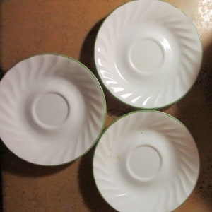 May include: Three white ceramic saucer plates with a spiral design and a green trim. The saucers are arranged on a brown surface, with a stack of similar plates visible in the background. The plates are designed to hold a teacup.