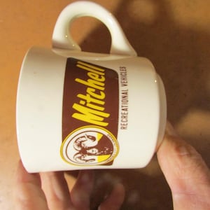 May include: Cream-colored ceramic mug with a curved handle. The mug features a brown rectangular label with the name "Mitchell" in yellow lettering. Below the name is a circular emblem with a ram's head and the words "Recreational Vehicles".