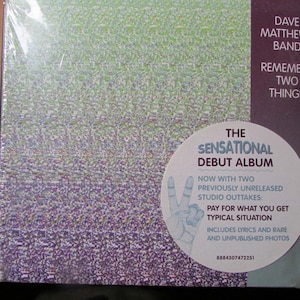 May include: A Dave Matthews Band Remember Two Things music album CD case with a green and purple patterned cover. A circular white sticker features the text The Sensational Debut Album, a peace sign graphic, and track titles.