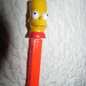 May include: A yellow Bart Simpson PEZ dispenser with a red neck and an orange dispenser body. The dispenser features Bart's face with large eyes and a mischievous expression. A collectible item.