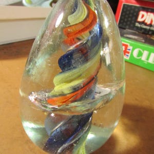 May include: A clear glass paperweight with a colorful, swirled design. The interior features a spiral of blue, yellow, and orange hues. The egg-shaped paperweight is a decorative item.
