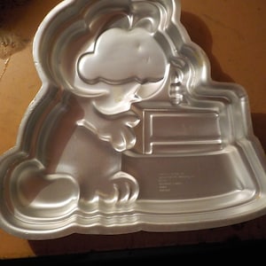 May include: A silver-colored metal cake pan in the shape of a cartoon dog holding a sign. The pan has a loop for hanging and is designed for baking a cake in a unique shape. The pan has text on the bottom.