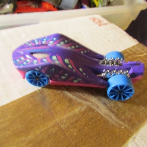 May include: A purple toy car with blue wheels and pink accents. The car has a unique design with a pattern of pink and green shapes. The wheels are a bright blue color, and the car appears to be made of plastic.