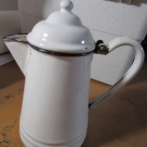 May include: White enamelware coffee pot with a black rim and handle. The pot has a lid with a small knob on top. The handle is attached to the side of the pot with a hinge. The pot is sitting on a patterned surface.