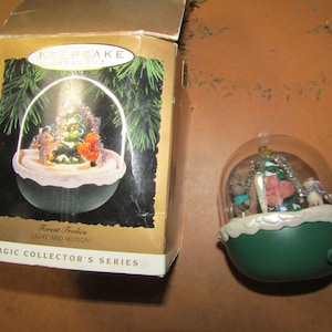 May include: A Christmas-themed snow globe with a green base and clear dome, featuring miniature figures around a small Christmas tree. The box next to it has a similar design with the text "Forest Frolic" and "Magic Collector's Series".