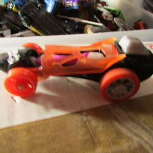 May include: An orange and black toy race car with red wheels. The car has a transparent orange body, revealing internal components. The wheels are a bright red color. The car is a toy vehicle.
