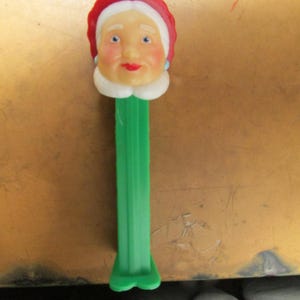 May include: A vintage PEZ dispenser featuring a Mrs. Claus head with a red hat, white hair, and a smiling face. The dispenser has a green stem and base. A collectible item.