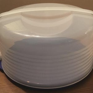 May include: A clear plastic cake carrier with a white handle and a blue base. The carrier has a domed lid with horizontal ridges and two white side clasps. The base is round and blue, providing a secure foundation for transporting baked goods.