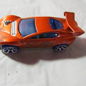 May include: Orange die-cast toy car with blue tinted windows and a rear spoiler. The toy car has blue wheels and a weathered appearance, suggesting it has been played with. The car is a miniature replica of a sports car.