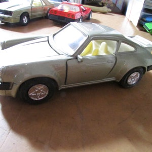 May include: A silver-colored toy car with black tires and a white interior. The car has a spoiler and star-shaped hubcaps. Other toy cars are visible in the background.