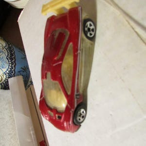 May include: A red toy car with a yellow spoiler. The car has black wheels and a clear windshield. The toy car is a miniature replica of a sports car.