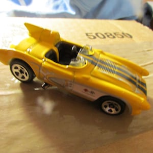 May include: Yellow toy race car with black and gray stripes on the hood. The car has a sleek design with a prominent fin and silver wheels. The car has the number 50850 printed on the side.