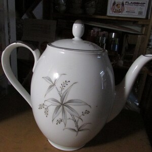 May include: White ceramic teapot with a silver rim and handle. The teapot features a floral design in gray and brown. The lid has a small, rounded knob. The teapot is on a wooden surface.
